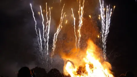 A group of people in the foreground watching a bonfire with fireworks illuminating the sky