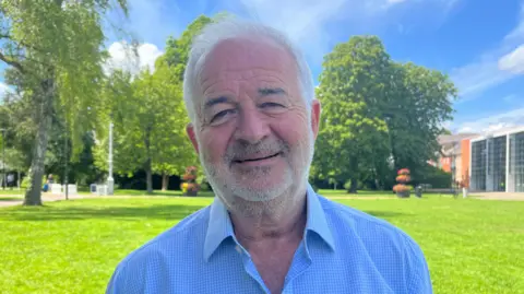 Tony Fisher/BBC A man with grey hair and short beard wearing a blue shirt. He is facing the camera standing in a park. There are trees and grass in the background as well as a building in the far right corner.