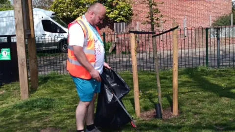 IRC A man in a hi-vis tabard and shorts is holding a litter-grabber and a black bin bag. He is in a grassy area by a tree.