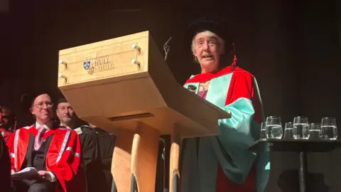 University of Hull Lesley Gracie stood making a speech behind a lectern. She is wearing a black cap and a red and turquoise gown.