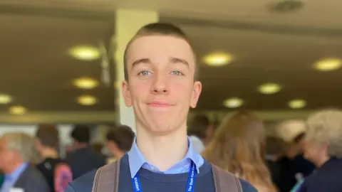 A boy with short hair smiling towards the camera, he's wearing a brown backpack and you can see the straps on his shoulders. He's also wearing a lanyard and a blue jumper.