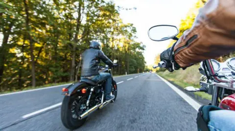 Two motorcyclists ride on a road through an area of forest.