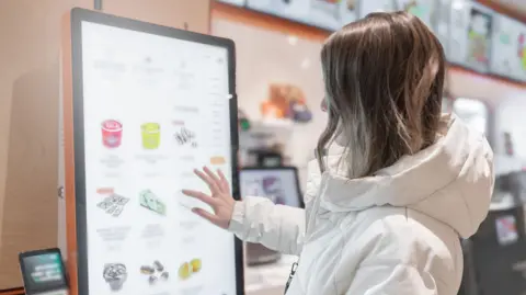 A woman touches the screen of a self-service terminal at a fast-food restaurant