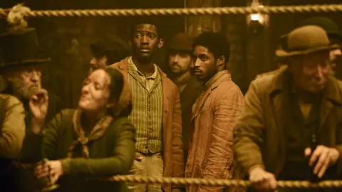 Malachi Kirby (left) and Francis Lovehall (right) in costume as their characters Hezekiah Moscow and Alec Munroe, looking into the distance at a boxing match