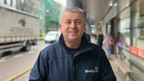 Kevin Hart wearing a dark LibertyBus jacket is standing at the bus station. A truck and other buildings line the street in the background with people to his right heading through the doors.

