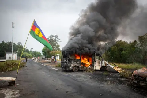 DELPHINE MAYEUR/AFP A Kanak flag waves next to a burning vehicle at an independantist roadblock at La Tamoa, in the commune of Paita, France's Pacific territory of New Caledonia on 19 May 2024