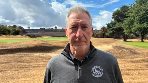 A man with grey hair and a dark grey fleece standing on a golf course with dry yellow grass and a couple of trees
