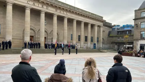 Four people look across the City Square, with their backs to the camera. Four flag bearers stand in front of Caird Hall steps where veterans are gathered for a memorial service.