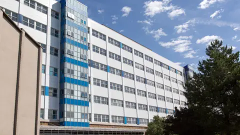 Getty Images A large white hospital building with horizontal rows of windows. One column of panelling has blue accents on. A tree can be seen on the left hand side, with white blue skies above.