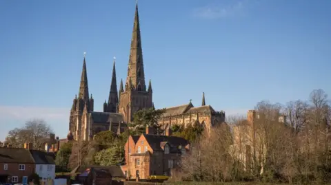 The large stone cathedral is captured from distance with buildings and trees in front of it. Its tall spire stands out in the bright sunshine.