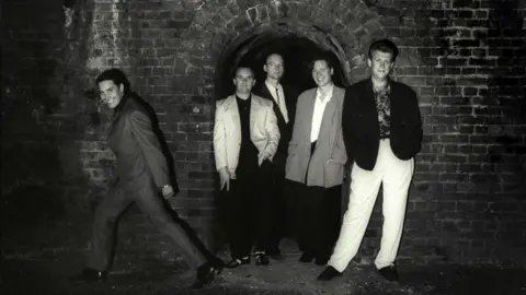 Getty Images A black and white image of the band Squeeze, the five male members of the band wear oversized suits and are posing in what looks like an underground tunnel with an archway behind them.