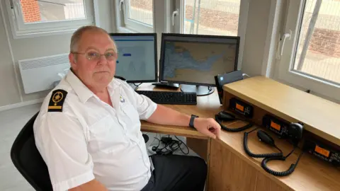 Simon Booth in his Coast Watch uniform of a white shirt and black trousers, at a desk with radios, in front of screens with maps and tide times on