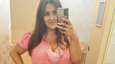 Melissa Ashcroft Melissa Ashcroft wearing a pink patterned dress taking a mirror selfie. She has straight brown hair and is smiling. 