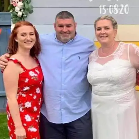 Summer Mace Three people are smiling at the camera with their arms around each other. A young woman on the left wears a red sun dress. The man in the middle wears a blue Oxford shirt and dark blue shorts and the woman on the right is in a white wedding dress. 