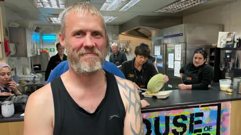 A man in a black vest with short light hair stands in front of a cafe kitchen area. He has a sleeve tattoo on his left arm. Three women behind the kitchen cafe counter eat, pour food into a container and look at the man being photographed.