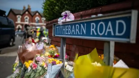 PA Media Bunches of flowers and teddies lined up against a wall next to a road sign reading Tithebarn Road.