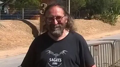 A man wearing a black t-shirt and glasses, he has thinning long black hair, a beard, and glasses. 