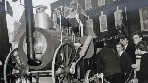 Family album A black and white photo of four teenage boys - mostly smiling - and standing next to a chain which has fenced off a replica of the engine, with a model of Father Christmas at the controls. Lloyds bank sign is in the background.