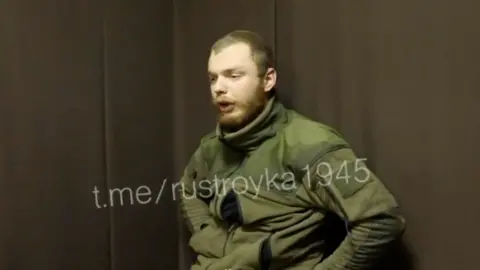 BBC A man believed to be James Scott Rhys Anderson, 22, a former British Army soldier captured while fighting for Ukraine, is seen wearing green military clothing and sat in front of a plain brown background.