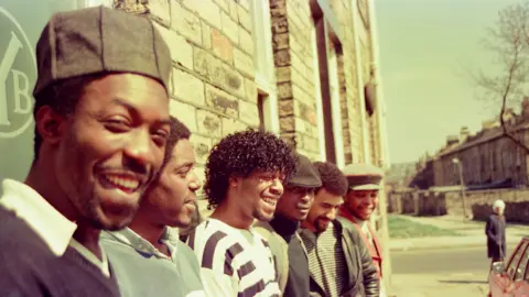 Six men smiling as they lean against the stone wall of a building.