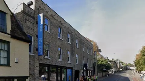 Google View of the front of Kettle's Yard gallery in Cambridge. It is a brick building with banners and signs on it showing its name. It is on a busy city street and there are a number of school children outside with a teacher, waiting at a pedestrian crossing