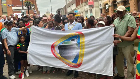 Kate Bradbrook/BBC Lots of people are stood in a street crowded around a white banner which has a logo on the front. The logo says Windrush Generations on it, and has the date of 1948 in the middle. A man who appears to be the Mayor, wearing a gold chain around his neck, stands in the middle.