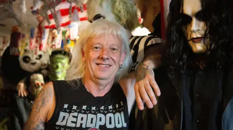A man with long white hair leaning against a life-sized prop with black hair and lipstick