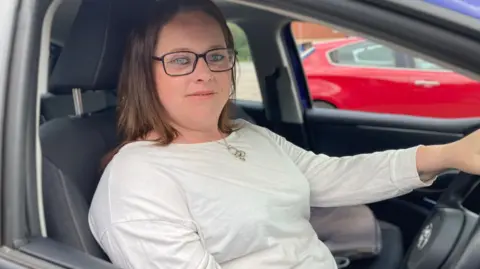 Simon Spark/BBC A bespectacled Lucy Bell sat in the driver's seat wearing a white top. She has brown shoulder-length hair.