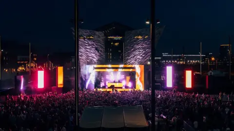 AVA, Photographer Hype A stage is lit up in the dark with yellow, orange and purple lights. The Titanic building can be seen in the background which is silver with wave like features along the walls. A large crowd can be seen in front of the stage.