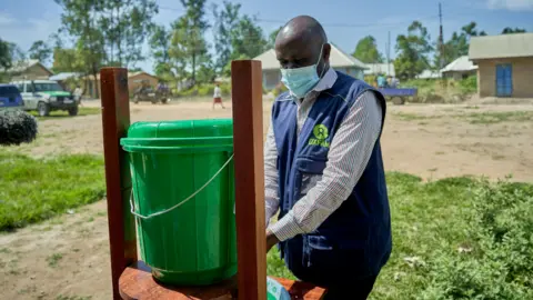 Azaria Muhongya / Oxfam A man with an Oxfam branded vest and a surgical mask on is washing his hands using a washing stand. He is in an area of Congo. It is a sunny day.