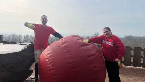 Elliott Webb and Tammy Gooding stand beside a giant red nose that is nearly as tool as Gooding. The pair are stood in red tops and yellow gloves.