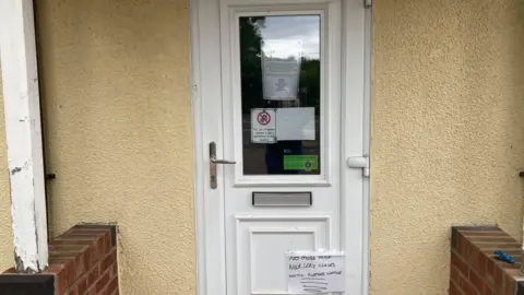 A white door on a yellow/beige wall. On the door are various signs. One that is visible, reads "No more milk. Nursery closed until further notice."