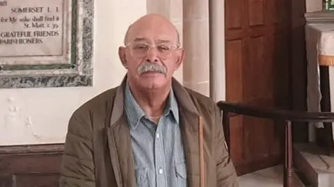 John Stockley wearing a grey shirt and brown anorak standing in front of a war memorial at the front of a church with the altar in the background.