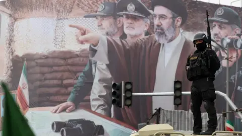 A member of the Iranian security forces stands guard in front of a billboard depicting the new Supreme Leader, Mojtaba Khamenei, and Iranian military and Revolutionary Guards commanders, during a rally to mark Quds Day at a square in Tehran, Iran (13 March 2026)