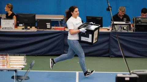 A woman with dark hair wearing a white T-shirt and jeans is running with a ballot box in 2017