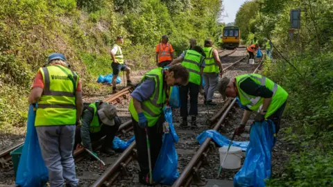 About a dozen male and female volunteers with his visibility vests and litter pickers are collecting waste from a rail line and placing it in blue refuse bags. The rails are surrounded by green shrubs. The sun is shining and a yellow train can be seen in the distance. 