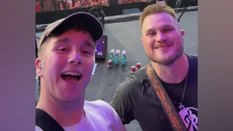 The image is a selfie of Paul with Zach Bryan. They are standing on a stage.