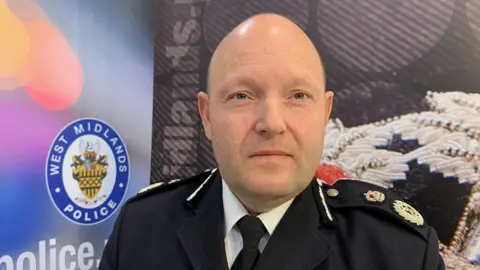 PA Media Chief Constable Craig Guildford wears his uniform and sits in front of a backdrop with the West Midlands Police badge on it. He is bald and has blue eyes, and a serious expression.