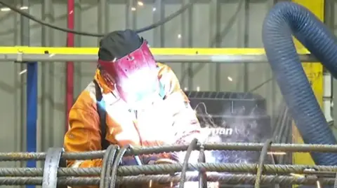 A worker at ArcelorMittal Kent Wire based at Chatham Docks. The worker is wearing an orange work suit and a face shield. 