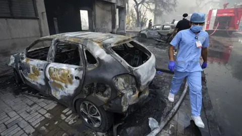 A man in medical scrubs walks beside a burnt out car
