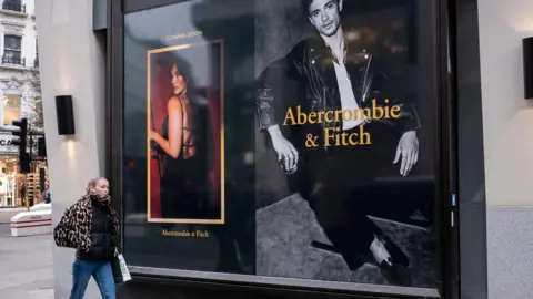 Getty Images A woman wearing jeans and a leopard print coat walking past a new Abercrombie & Fitch store in central London, with a window display featuring a male model photographed in black and white wearing a white T-shirt and a leather jacket, and a female model in a black dress with straps across the back, looking over her shoulder against a red background.