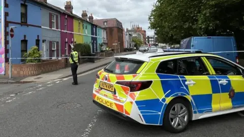 A row of brightly coloured houses with a floral mural at the gable end with the word "Gloucester" picked out in blue and yellow. The road is cordoned off and a police car is blocking traffic. A police officer stands next to police tape. 
