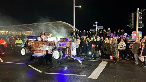 BBC An illuminated cart Weston's Carnival, with crowds looking on. The procession has a man in a glittery red hat and woman sat close by.