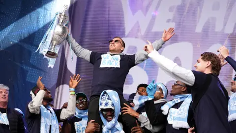 PA Media Jamie Allen is on a team-mate's shoulders, holding the Championship trophy aloft on stage. Lots of other players are in the background, smiling