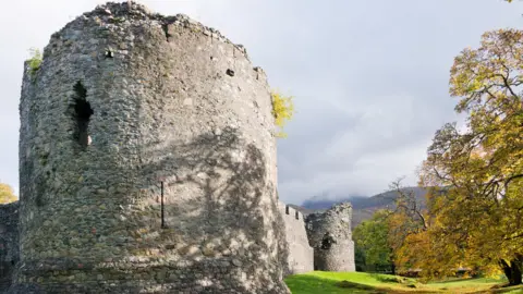 The image show a corner tower at Old Inverlochy castle. Shadows from trees are being cast on to the stonework. The picture shows an autumnal scene with leaves turning yellow and orange. further parts of the castle's wall and another corner tower can be seen in the background along with mist covered mountains. 