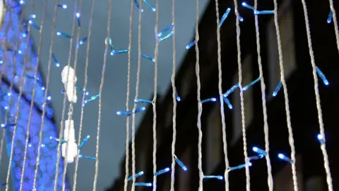 Columns of strings of Christmas lights, which are illuminated blue. A larger blue light installation is illuminated but out of focus in the background.