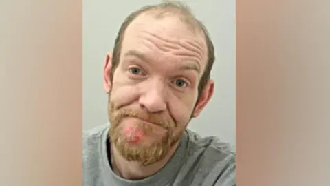 Craig Cotton's mugshot. He has receding short brown hair with tufts on top and a gingerish beard. 