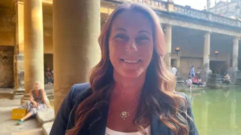 Penny is a woman with long curly auburn hair. She is wearing a golden crab necklace and a blue blazer and is smiling at the camera. She stands in front of the thermal bath. 