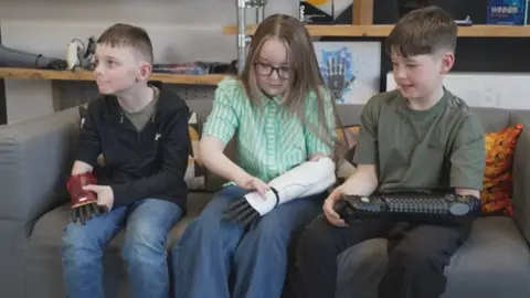 Three children, two boys with a girl in the middle, sitting on a sofa, all aged about nine and wearing their new bionic arms. They are looking at them and feeling what's they're like to wear