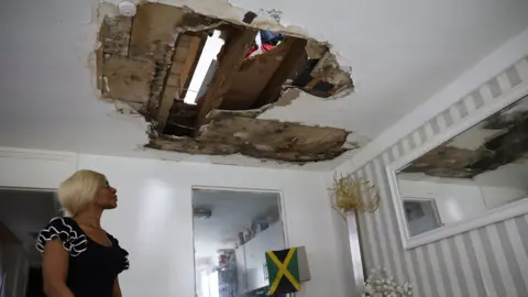 Sacha Berko, a woman with blonde hair standing on the left looking upwards at the ceiling where a hole showing the room above has appeared.